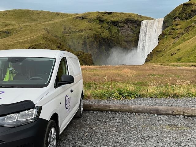 Camper van parked near a massive waterfall, with the spray visible in the air and lush green cliffs surrounding the area in Iceland.