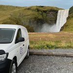 Camper van parked near a massive waterfall, with the spray visible in the air and lush green cliffs surrounding the area in Iceland.