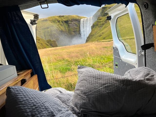 View from inside a camper van showing a cozy bed setup with the door open to a stunning view of a large waterfall and lush green cliffs in Iceland.