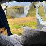 View from inside a camper van showing a cozy bed setup with the door open to a stunning view of a large waterfall and lush green cliffs in Iceland.