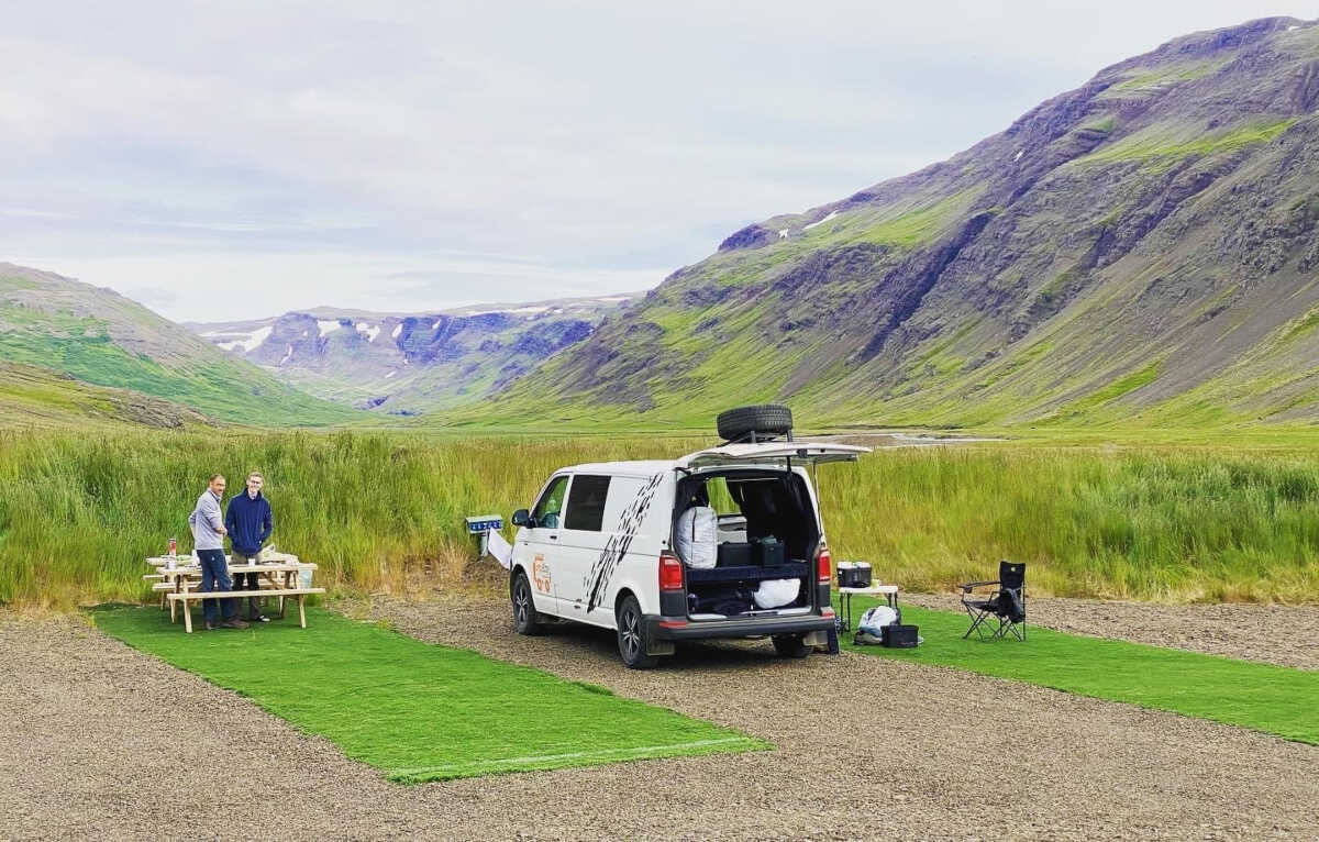 Two travelers enjoying a meal at a picnic table beside a white Camp Easy camper van in a picturesque Icelandic valley. The van's trunk is open, displaying camping gear.