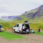 Two travelers enjoying a meal at a picnic table beside a white Camp Easy camper van in a picturesque Icelandic valley. The van's trunk is open, displaying camping gear.