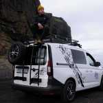 A man sits atop a roof rack of a white camper van, wearing a yellow beanie and a dark jacket. The van is parked on a gravel surface near a towering dark cliff, creating a moody and dramatic atmosphere.