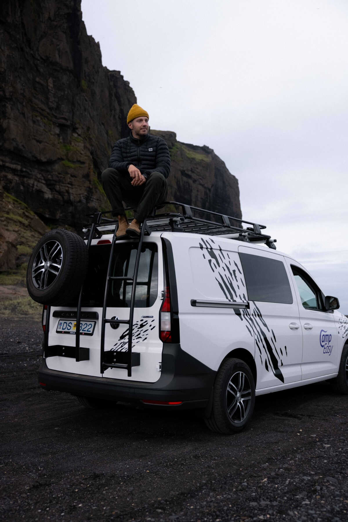 A man sits atop a roof rack of a white camper van, wearing a yellow beanie and a dark jacket. The van is parked on a gravel surface near a towering dark cliff, creating a moody and dramatic atmosphere.