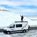 A person stands on the roof rack of a white camper van, parked on a snowy road surrounded by vast snowy mountains. The person is dressed in black, waving happily under a clear blue sky.