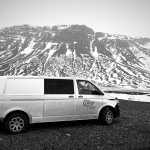 A white camper van parked on a gravel surface in front of a large, snow-patched mountain. The surroundings appear cold and desolate, reflecting a stark yet majestic winter landscape