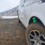 Close-up of a white CampEasy campervan's muddy tire and side logo, with a scenic mountainous backdrop in Iceland, ideal for adventurous road trips.