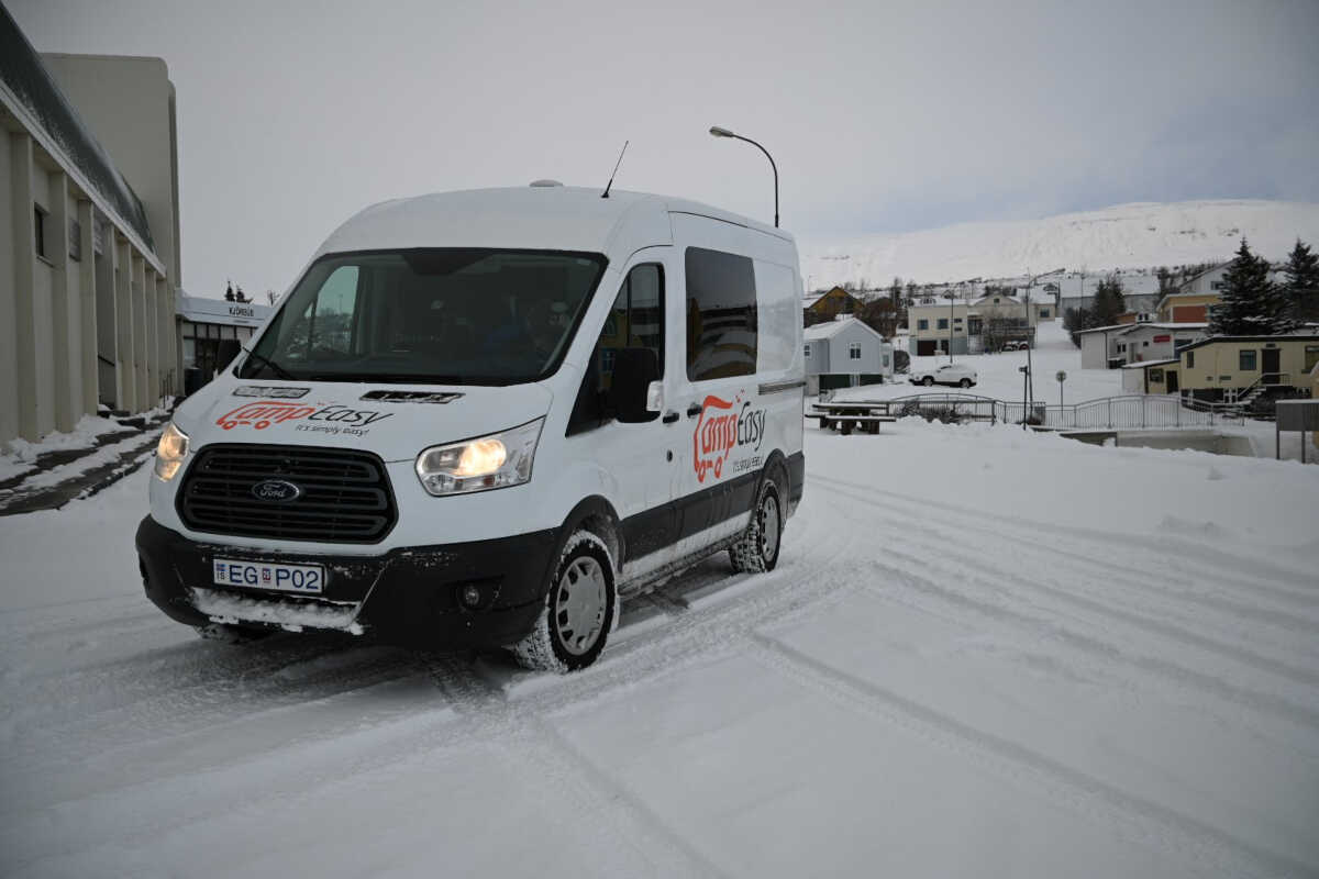 CampEasy campervan navigating through a snow-covered street in a small Icelandic town, with quaint houses and a snow-capped mountain in the background during a gray winter day