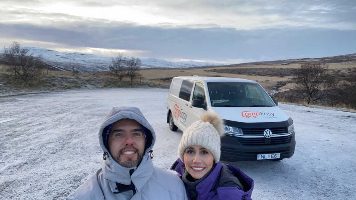 Smiling couple in winter clothing taking a selfie with a Camp Easy campervan parked in a snowy landscape, with rolling hills and a frosty terrain in the background in Iceland.