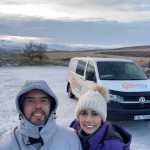 Smiling couple in winter clothing taking a selfie with a Camp Easy campervan parked in a snowy landscape, with rolling hills and a frosty terrain in the background in Iceland.