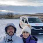 Smiling couple in winter clothing taking a selfie with a Camp Easy campervan parked in a snowy landscape, with rolling hills and a frosty terrain in the background in Iceland.