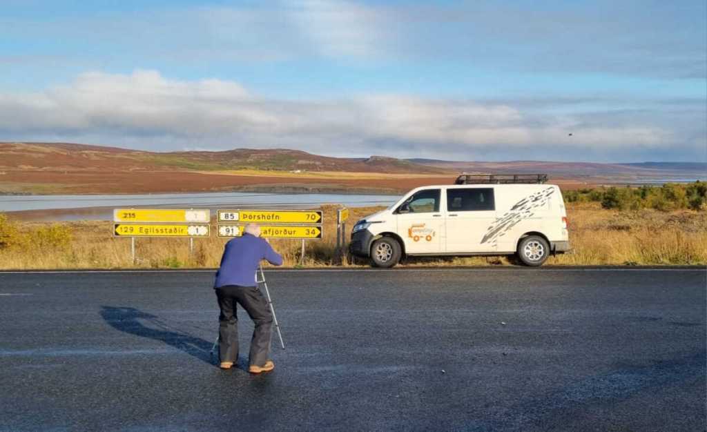 A person taking a picture on a road in front of a parked campervan near a directional signpost in a rural area. The signpost indicates distances to various destinations such as Porshofn and Egilsstadir. The background features a wide landscape with low hills and a large body of water under a clear, sunny sky.