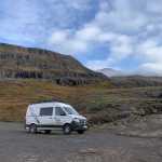 A campervan parked on a rocky trail with a dramatic backdrop of steep, layered mountains and autumn-colored vegetation under a partly cloudy sky in Iceland.