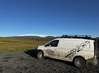A campervan parked on a gravel path with a distant view of a snow-capped mountain in Iceland, showcasing an adventurous travel experience in a pristine landscape.