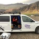 Woman sitting in an open Camp Easy campervan, surrounded by camping gear, with dramatic mountainous scenery in the background in Iceland.