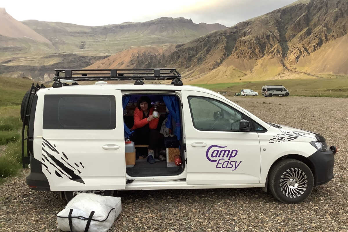 Woman sitting in an open Camp Easy campervan, surrounded by camping gear, with dramatic mountainous scenery in the background in Iceland.