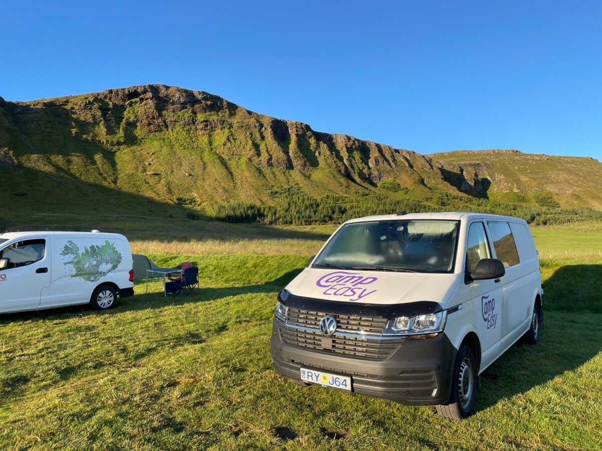 Two white campervans parked on lush green grass with folding chairs set up, overlooking a stunning cliff in Iceland.