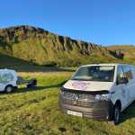 Two white campervans parked on lush green grass with folding chairs set up, overlooking a stunning cliff in Iceland.