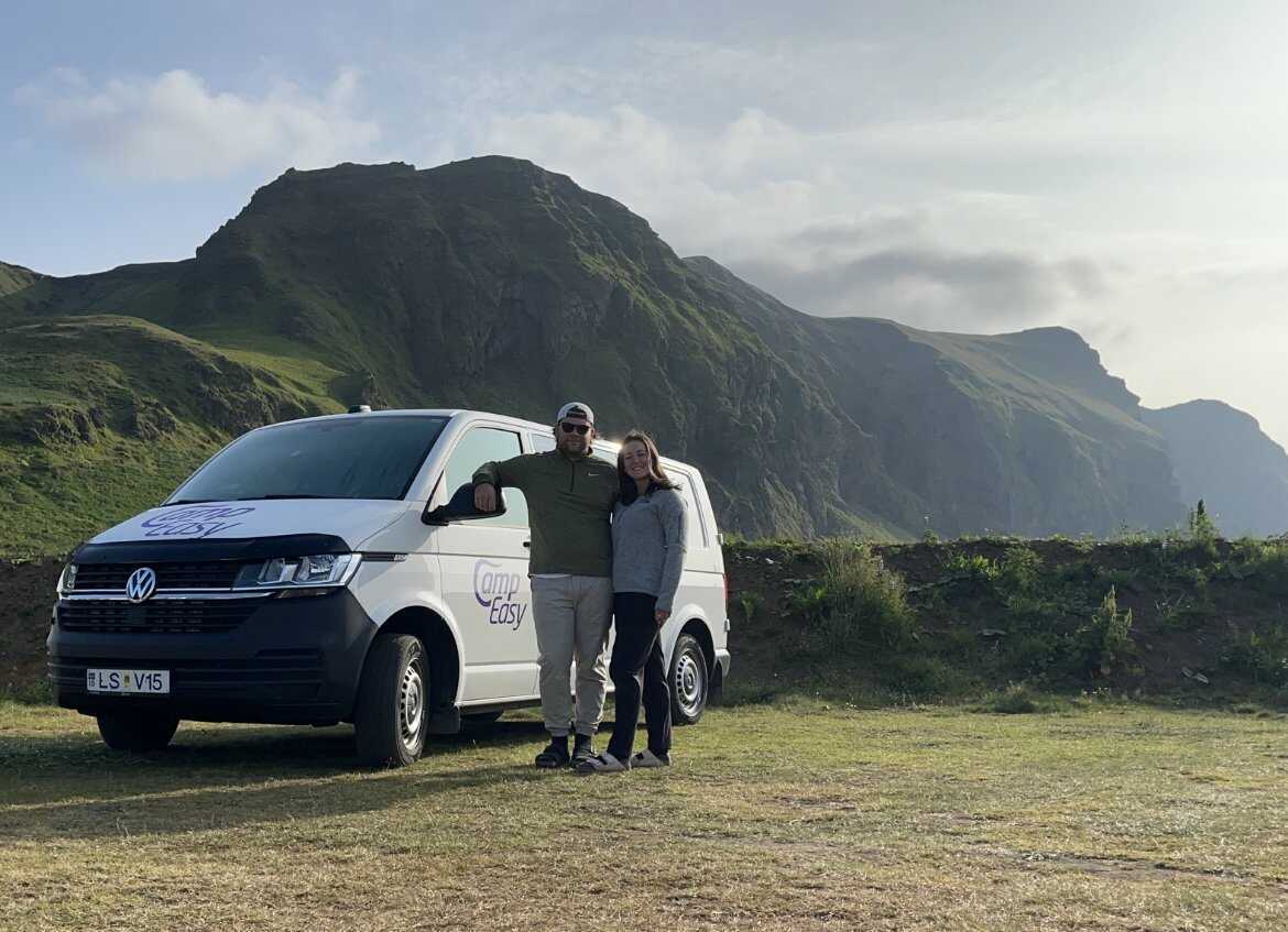Happy couple standing beside a white campervan with scenic green mountains in the background in Iceland.