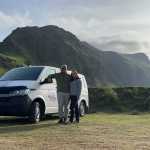 Happy couple standing beside a white campervan with scenic green mountains in the background in Iceland.