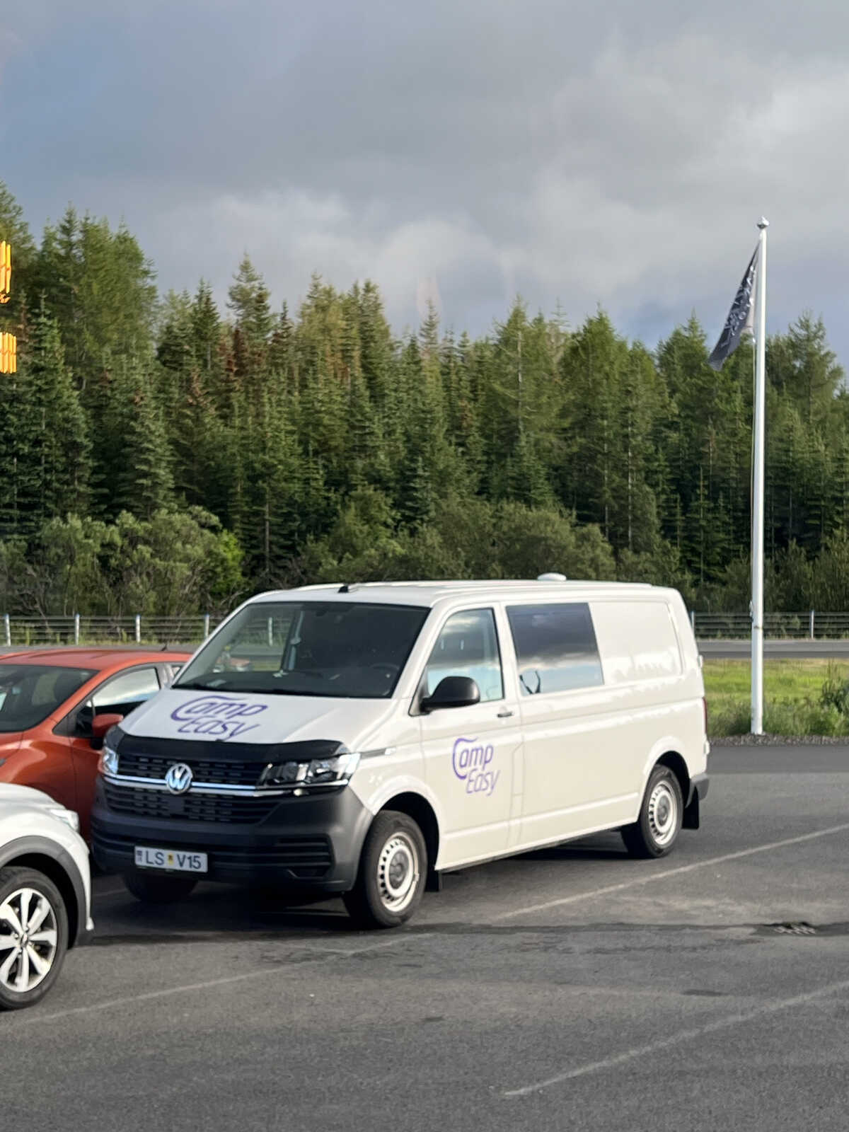 A white campervan parked in a lot, with a forest of green trees in the background and a flagpole nearby, under a cloudy sky in Iceland.
