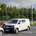 A white campervan parked in a lot, with a forest of green trees in the background and a flagpole nearby, under a cloudy sky in Iceland.