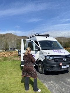 A person photographing a white CampEasy van equipped with a roof rack, parked in a scenic outdoor area with sparse trees under a clear blue sky.