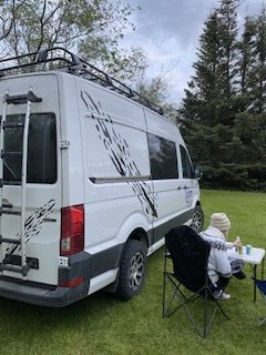 Person relaxing in a folding chair beside a decorated white campervan, surrounded by lush greenery, depicting a peaceful camping experience.