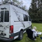 Person relaxing in a folding chair beside a decorated white campervan, surrounded by lush greenery, depicting a peaceful camping experience.