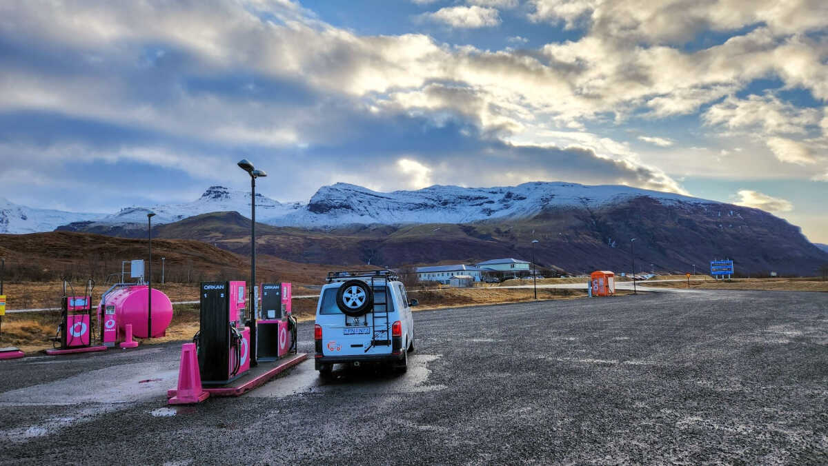 CampEasy campervan refueling at a vibrant pink gas station in a scenic Icelandic setting with snowy mountains in the background, under a partly cloudy sky.