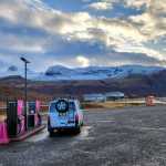 CampEasy campervan refueling at a vibrant pink gas station in a scenic Icelandic setting with snowy mountains in the background, under a partly cloudy sky.