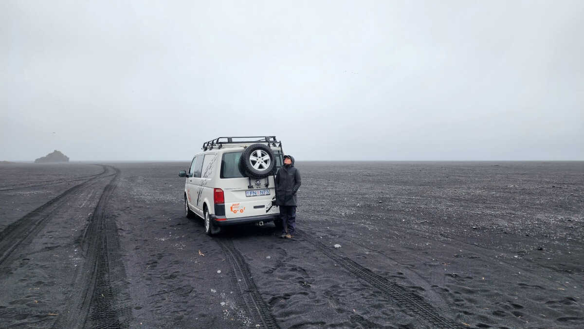 CampEasy campervan and a person standing on a vast volcanic black sand beach in Iceland, under a foggy sky, emphasizing the vehicle's capability to explore unique and remote landscapes.