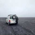 CampEasy campervan and a person standing on a vast volcanic black sand beach in Iceland, under a foggy sky, emphasizing the vehicle's capability to explore unique and remote landscapes.