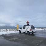 Person standing with arms outstretched on the roof of a CampEasy campervan parked by a coastal area in Iceland, with a vast ocean and snow-capped mountains in the background, under a cloudy sky.