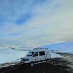 A white campervan parked on a roadside during winter, showcasing expansive snowy mountain landscapes under a clear blue sky in Iceland.