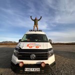 Joyful man standing with arms raised on top of a CampEasy campervan, set against a vast Icelandic landscape under a clear blue sky.