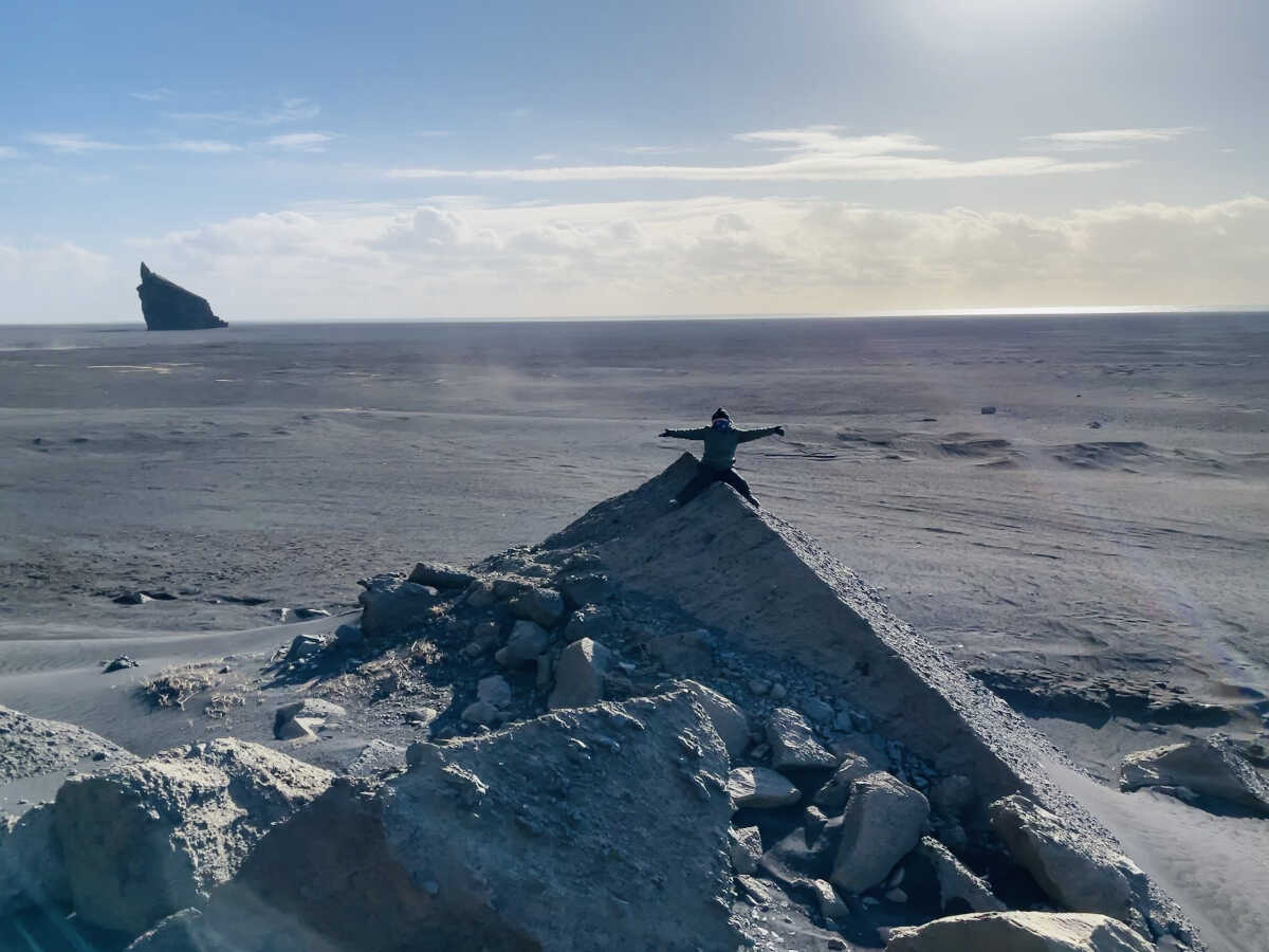 Person sitting atop a rocky outcrop with arms outstretched, overlooking the vast sandy shores of Iceland with a dramatic rock formation visible in the distance under a bright, clear sky.