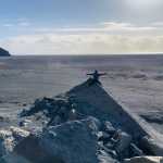 Person sitting atop a rocky outcrop with arms outstretched, overlooking the vast sandy shores of Iceland with a dramatic rock formation visible in the distance under a bright, clear sky.