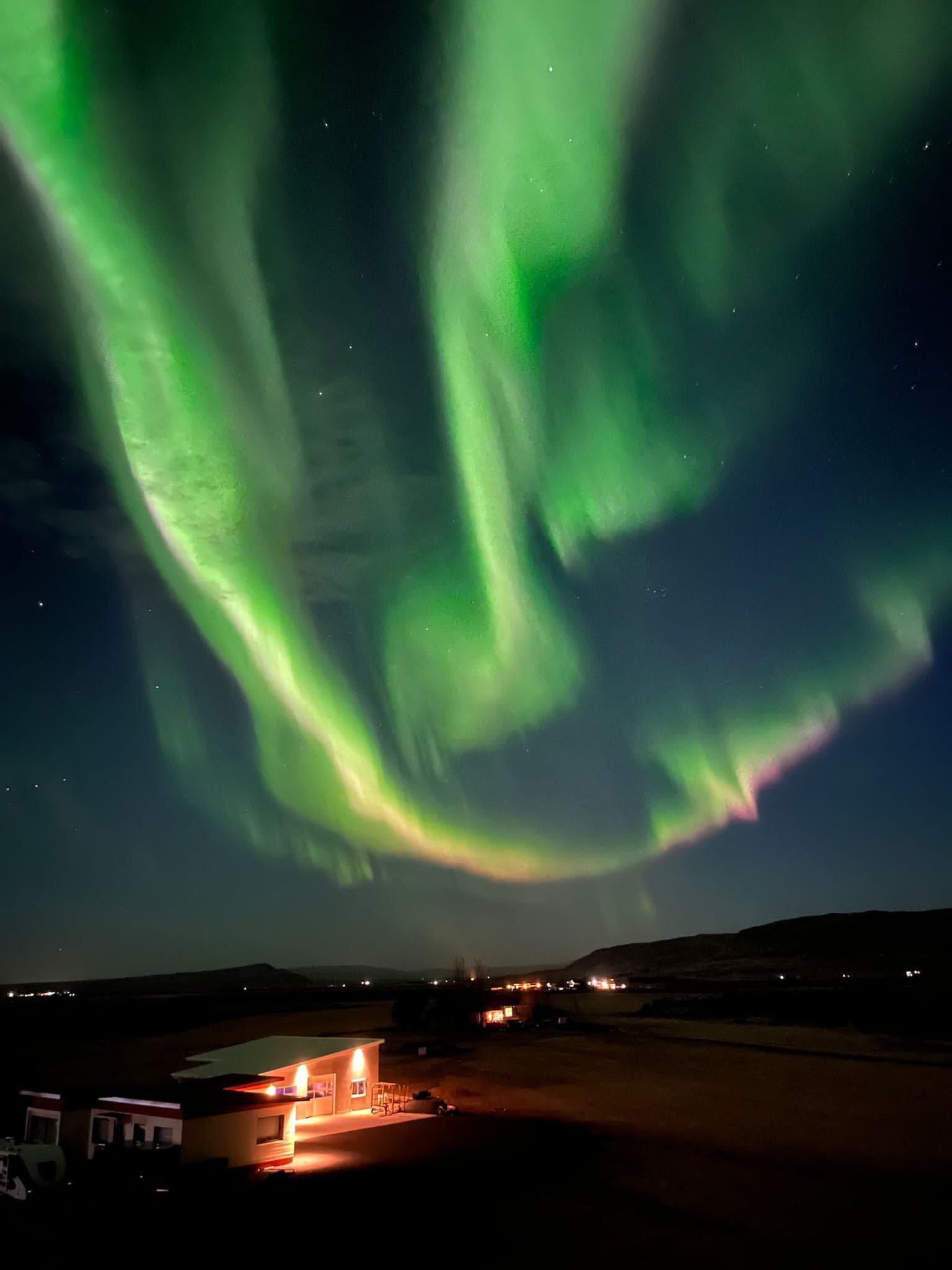 Dramatic display of the Northern Lights in vibrant green swirls above a lit building and campervans in a rural Icelandic setting, emphasizing the enchanting night skies unique to the region.