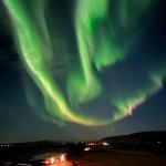 Dramatic display of the Northern Lights in vibrant green swirls above a lit building and campervans in a rural Icelandic setting, emphasizing the enchanting night skies unique to the region.