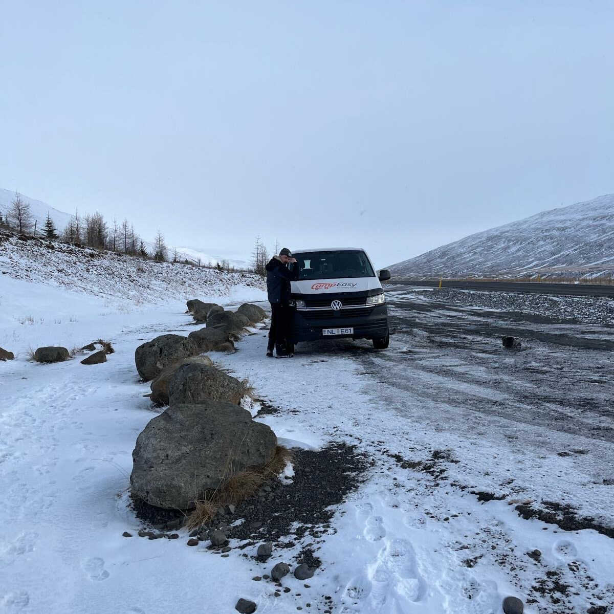 A person in dark winter clothing is taking a photo of a scenic view in Iceland, leaning on a CampEasy branded campervan parked along a snowy roadside surrounded by barren hills.