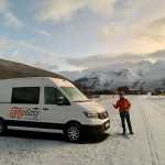 A person in an orange jacket stands next to a CampEasy campervan on a snow-covered road in Iceland, with majestic snow-capped mountains in the background during a bright winter day.