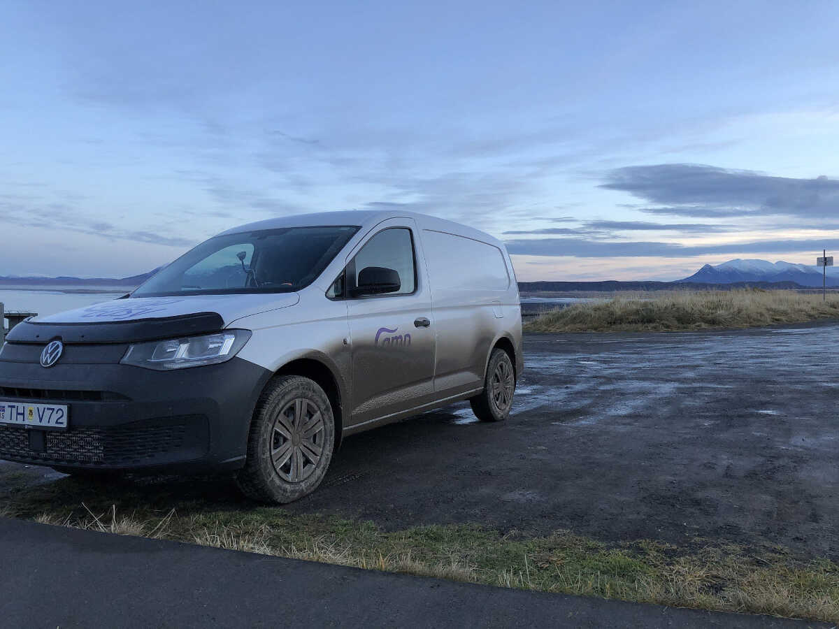 A camper van parked by a coastline with a clear view of distant mountains under a soft morning sky in Iceland.