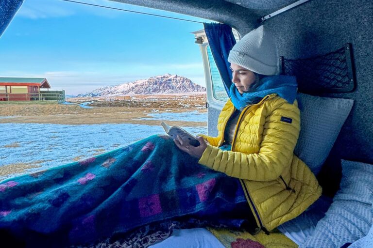 A woman covered with a blanket lies on a bed in a camper van and reads a book with the rear door open, through which the Icelandic landscape can be seen.