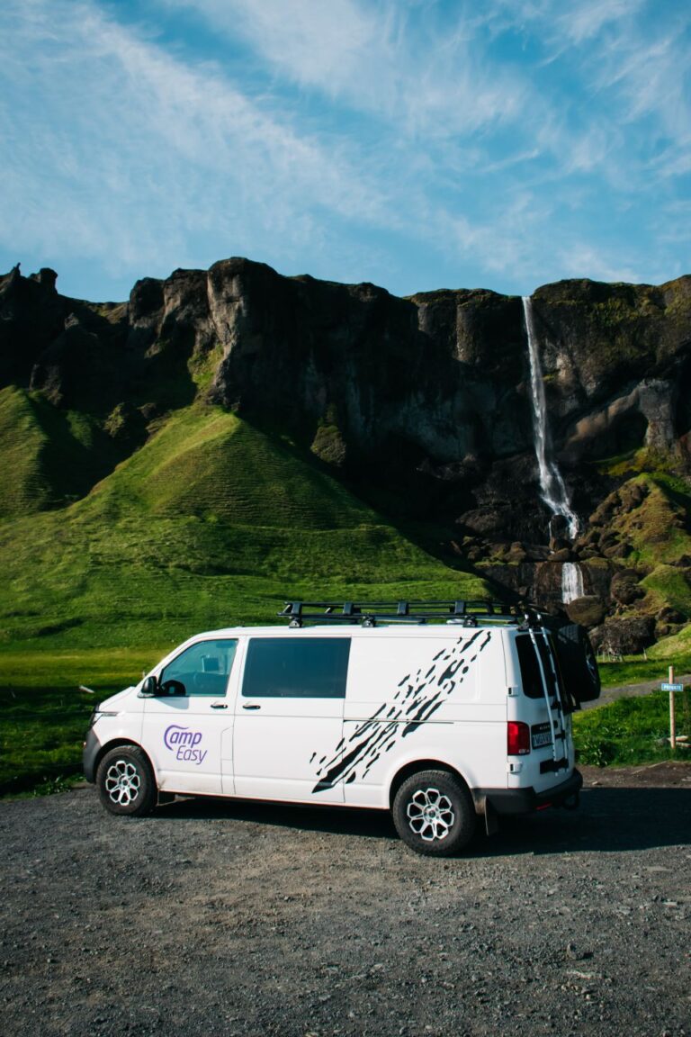 A campervan parked in a parking lot near a waterfall and mountains covered with green moss.