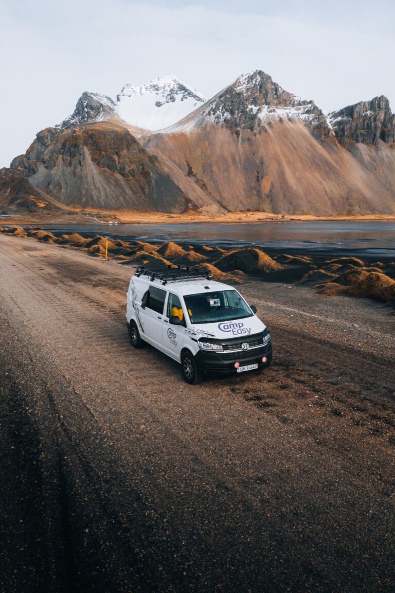 A campervan parked on a gravel road near a vast black sand beach and huge mountains