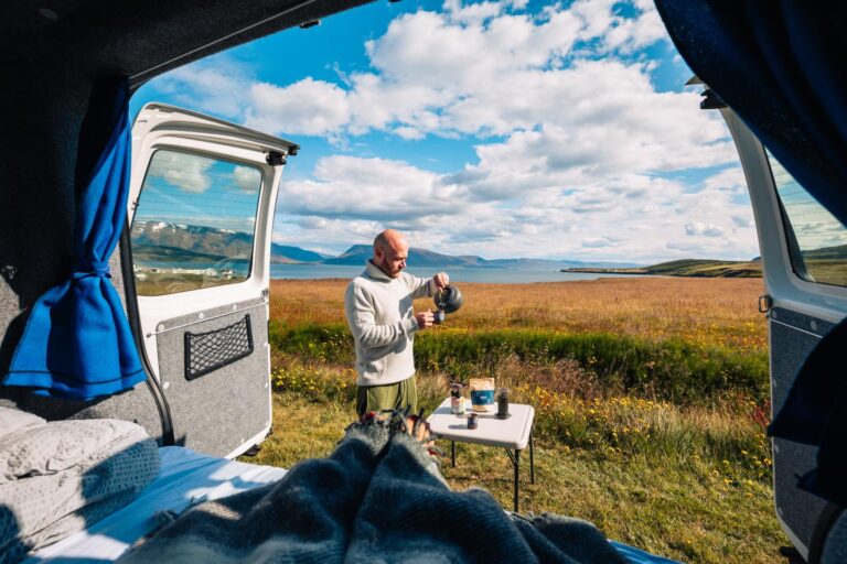 A man pours water into a cup while standing next to a tourist table set up near meadows overlooking the fjord.
