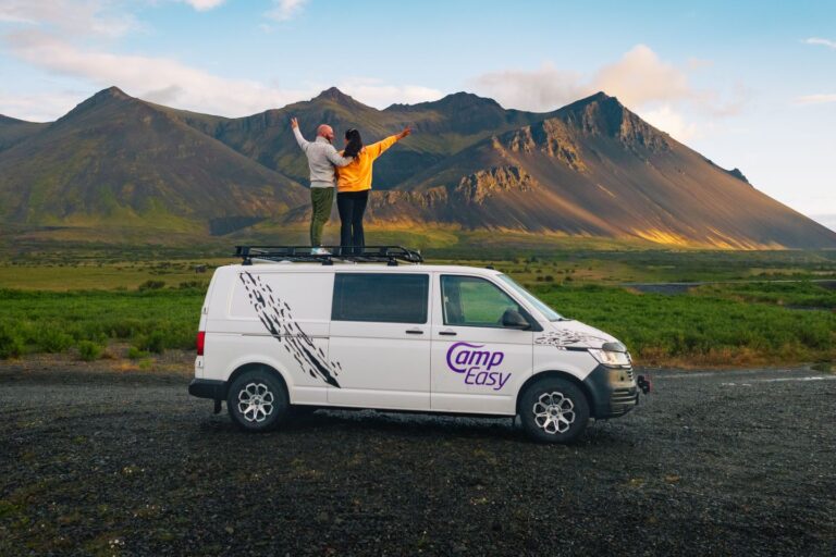 The couple stands on the roof rack of the campervan with their arms raised against the backdrop of huge mountains.