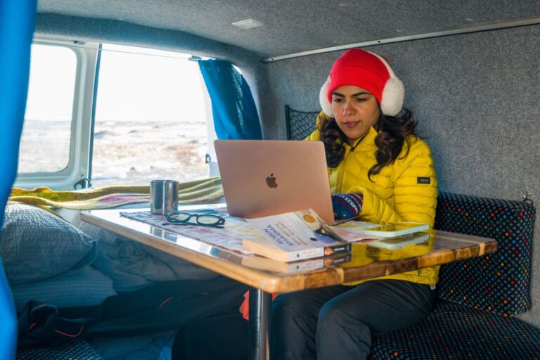A woman in a yellow jacket is sitting inside a camper van at a table, working on her laptop.