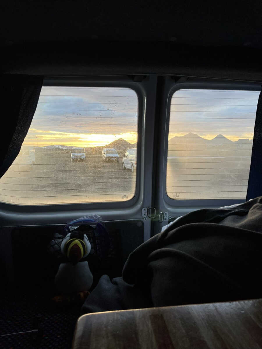 View from inside a campervan showing a sunset over distant Icelandic mountains through the window, with plush toys and a cozy blanket inside.